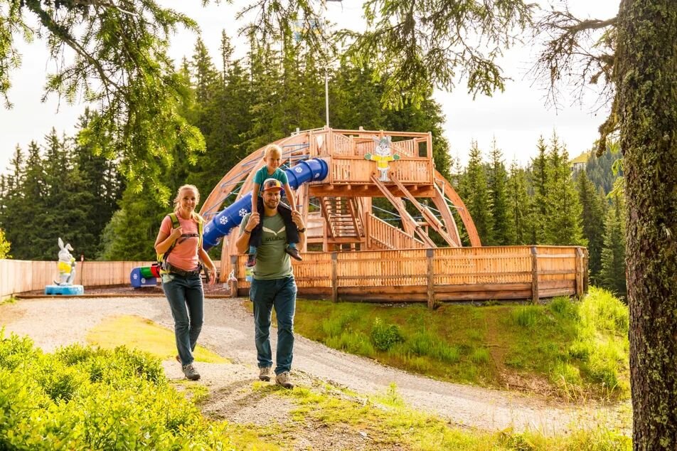 Familie mit Kind auf Schultern geht am Spielplatz vorbei