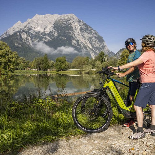 Zwei Radfahrer mit Helmen neben einem limettenfarbenen E-Bike am See.