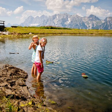 Zwei Mädchen stehen im flachen Bergsee am Grasufer.