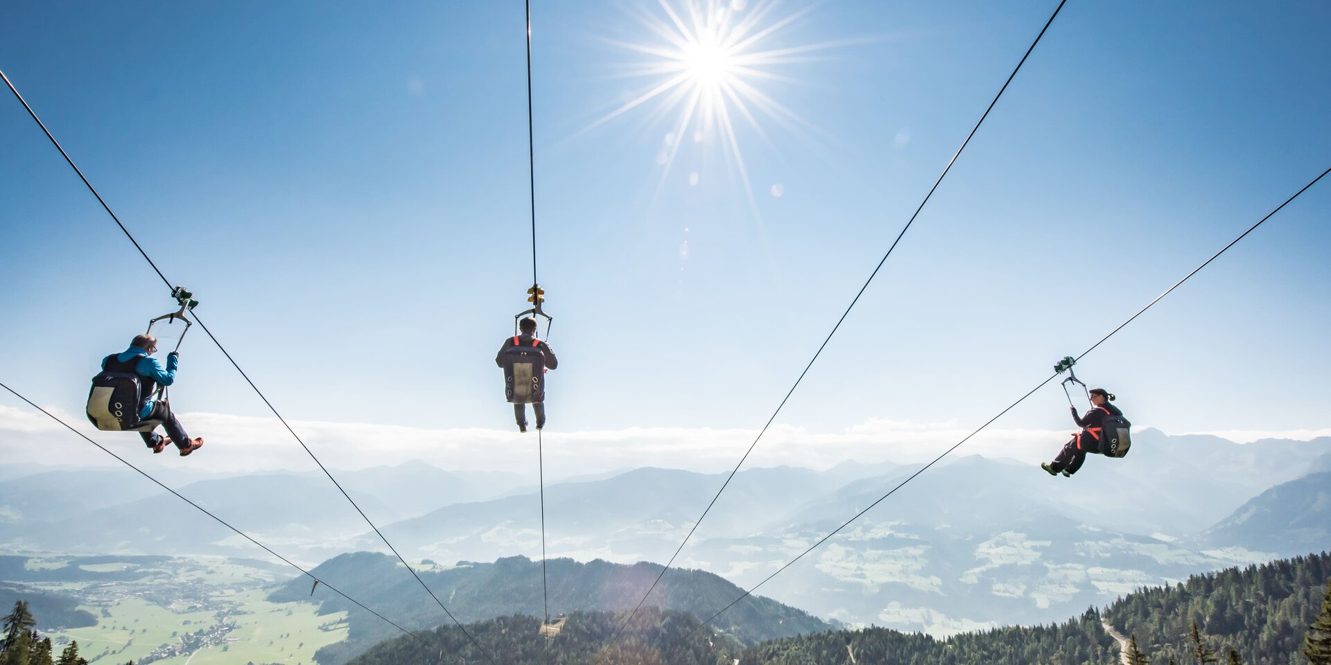 Drei Personen ziplinen über Berglandschaft bei Sonnenschein