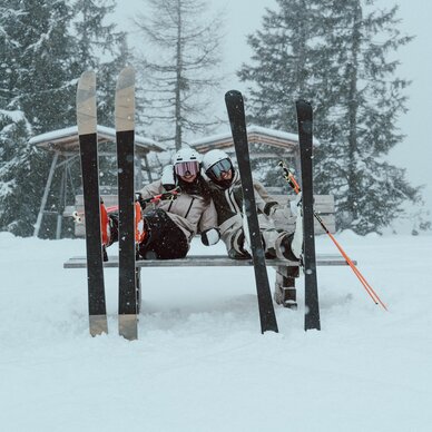 Zwei Skifahrer sitzen auf einer Bank im Schnee, Skier in der Nähe.