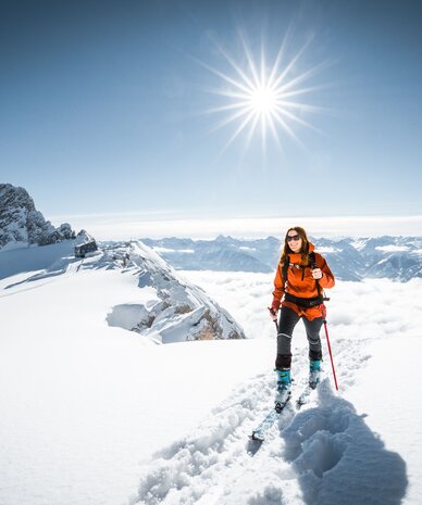 Skifahrer(in) in orangener Jacke auf verschneiter Bergkuppe unter strahlender Sonne