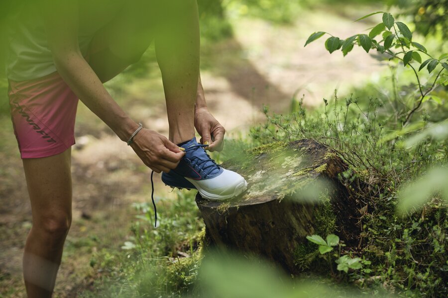 Person bindet blauen Wanderschuh an einem Baumstumpf im Wald | © RAPHAELGABAUER.COM
