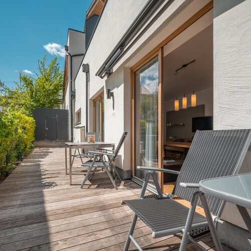 Holzterrasse mit Sitzgelegenheiten neben weißem Wohngebäude | © TIEBER