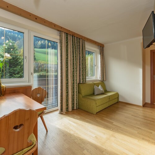 Heller Wohnbereich mit großen Fenstern, grünem Sofa und Esstisch aus Holz.