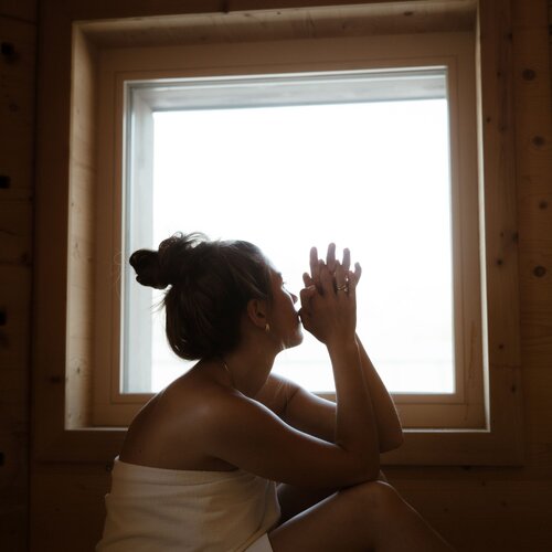 Frau im Handtuch sitzt am Fenster in einem Holzzimmer, Hände gefaltet