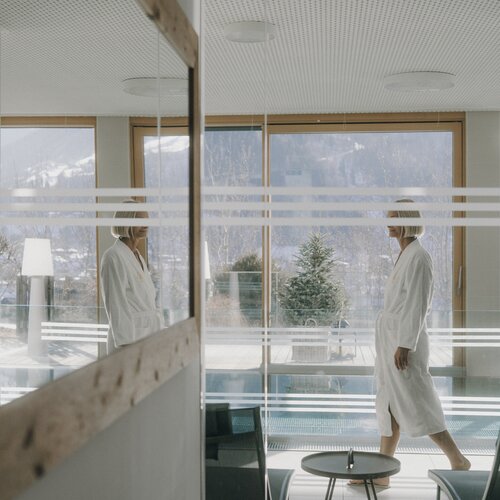 Zwei Personen in weißen Bademänteln in einer gläsernen Lobby.