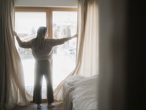 Woman standing by large window, drawing back beige curtains