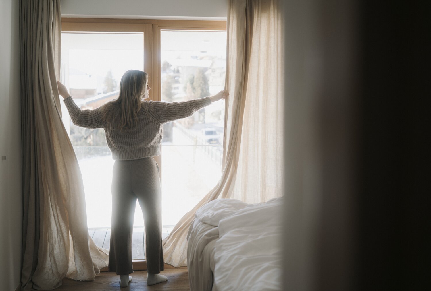 Woman standing by large window, drawing back beige curtains