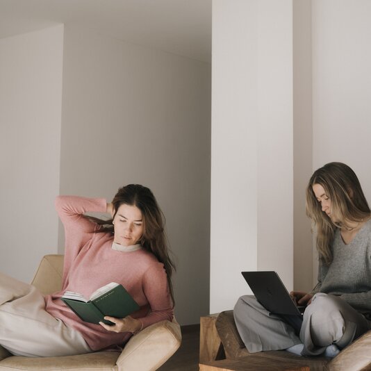 Two women in a modern living room; one reads, another works.