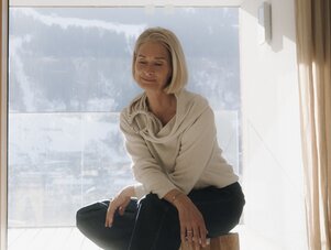 Older woman sits on wooden stool by window, smiling.